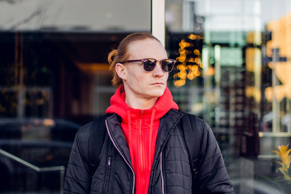 uomo che indossa felpa con cappuccio rossa con zip sotto giacca di pelle nera con zip in piedi accanto a una parete di vetro trasparente