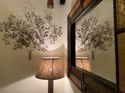 A decorative wall arrangement featuring metal branches with leaves adorns the wall above a warm-colored floor lamp. The lamp casts a soft glow, creating intricate shadows of the branches on the wall. A large ornate mirror is positioned to the right, reflecting the scene.