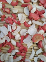 Slices of red and white vegetables are spread across a bamboo mat, likely for drying or preparation. The pieces vary in size and shape, with some displaying a circular form and others more irregular.