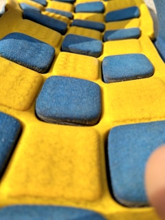 A close-up view of a textured surface featuring a pattern of alternating blue and yellow squares. The surface appears to be made of a soft, rubbery material, with the blue squares slightly raised above the yellow background.