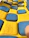 A close-up view of a textured surface featuring a pattern of alternating blue and yellow squares. The surface appears to be made of a soft, rubbery material, with the blue squares slightly raised above the yellow background.
