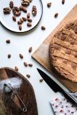 A rustic homemade pie with a lattice crust sits on a wooden board. Nearby, a small sieve rests on another wooden board sprinkled with powder, possibly flour or sugar. A knife lies next to the pie, and a floral napkin adds a touch of color. A plate holds a mix of walnuts and almonds, with additional almonds scattered across the table.
