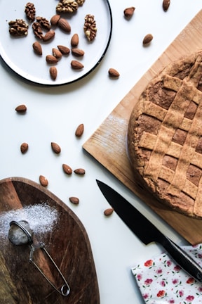 A rustic homemade pie with a lattice crust sits on a wooden board. Nearby, a small sieve rests on another wooden board sprinkled with powder, possibly flour or sugar. A knife lies next to the pie, and a floral napkin adds a touch of color. A plate holds a mix of walnuts and almonds, with additional almonds scattered across the table.