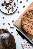 A rustic homemade pie with a lattice crust sits on a wooden board. Nearby, a small sieve rests on another wooden board sprinkled with powder, possibly flour or sugar. A knife lies next to the pie, and a floral napkin adds a touch of color. A plate holds a mix of walnuts and almonds, with additional almonds scattered across the table.