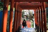 A series of bright red torii gates form a pathway down a stone stairway, with a person in a dark coat walking through them. The gates are adorned with black Japanese characters. Trees and a traditional Japanese building are visible in the background.