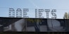 Large, bold letters spelling out 'DOE IETS' are prominently displayed, constructed from a grid-like metal structure. Below the letters is a photographic mural with various images, including graffiti and geometric architectural elements, cast in deep shadows. The sky is clear with a faint contrail visible.