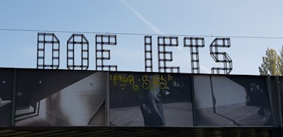 Large, bold letters spelling out 'DOE IETS' are prominently displayed, constructed from a grid-like metal structure. Below the letters is a photographic mural with various images, including graffiti and geometric architectural elements, cast in deep shadows. The sky is clear with a faint contrail visible.