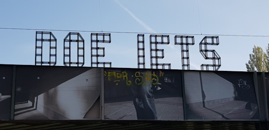 Large, bold letters spelling out 'DOE IETS' are prominently displayed, constructed from a grid-like metal structure. Below the letters is a photographic mural with various images, including graffiti and geometric architectural elements, cast in deep shadows. The sky is clear with a faint contrail visible.