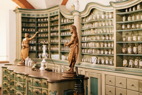 An ornate vintage apothecary filled with numerous shelves containing glass jars and bottles, each labeled. Two detailed statues are situated prominently on the wooden counter, alongside other classical vessels and scales, giving an old-world charm to the room.