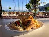 A plate of sandwiches and fries is placed on an outdoor table overlooking a scenic view of the sea. In the background, there are chairs and tables under a patio, and a glass of white wine is positioned next to the plate. Trees and sailboat masts are visible near the shoreline.