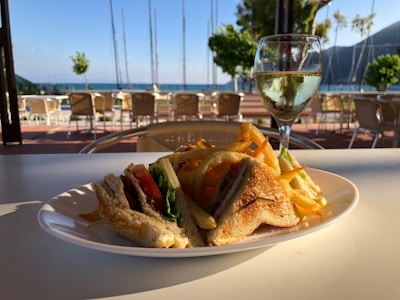 A plate of sandwiches and fries is placed on an outdoor table overlooking a scenic view of the sea. In the background, there are chairs and tables under a patio, and a glass of white wine is positioned next to the plate. Trees and sailboat masts are visible near the shoreline.
