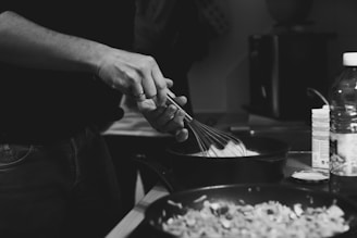A person is using a whisk to stir ingredients in a frying pan, focusing on cooking. Nearby, there are other cooking materials including a bottle and boxes.