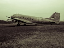 A vintage propeller airplane, marked with 'Indonesian Airways' and 'RI-001', is parked on an open grass field. The plane appears to be old and possibly not in service, with visible wear and a muted color palette.