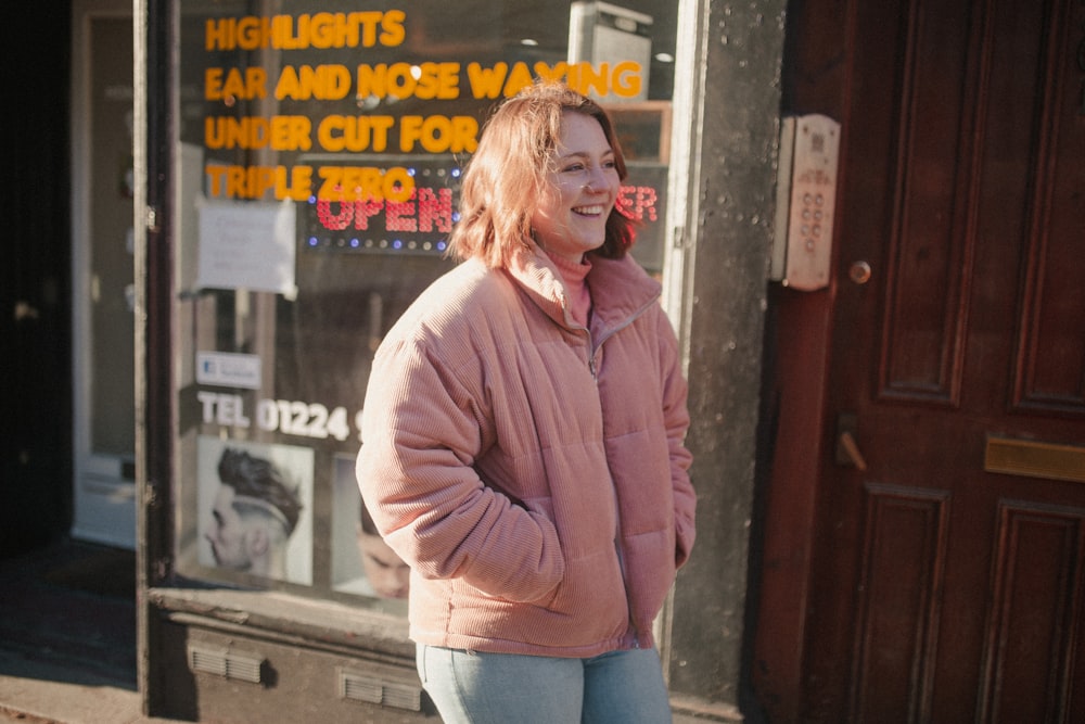 smiling woman wearing pink zip-up jacket standing near brown wooden door