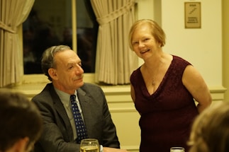 A man in a suit and tie sits at a table with a glass of water in front of him, while a woman in a sleeveless burgundy dress stands beside him, smiling. They are in a warmly lit room with cream-colored curtains and walls.