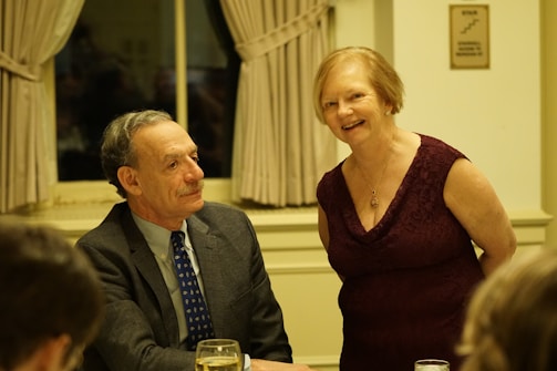 A man in a suit and tie sits at a table with a glass of water in front of him, while a woman in a sleeveless burgundy dress stands beside him, smiling. They are in a warmly lit room with cream-colored curtains and walls.