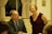 A man in a suit and tie sits at a table with a glass of water in front of him, while a woman in a sleeveless burgundy dress stands beside him, smiling. They are in a warmly lit room with cream-colored curtains and walls.