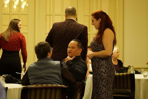 A group of people are socializing in a formal indoor setting. One woman with long red hair, wearing a patterned dress, is interacting with a man seated at a table, who is speaking expressively. Other people are gathered around, some seated and some standing. The ambiance suggests a gathering or event with a formal or semi-formal atmosphere.