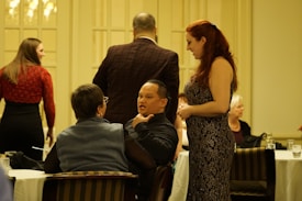 A group of people are socializing in a formal indoor setting. One woman with long red hair, wearing a patterned dress, is interacting with a man seated at a table, who is speaking expressively. Other people are gathered around, some seated and some standing. The ambiance suggests a gathering or event with a formal or semi-formal atmosphere.