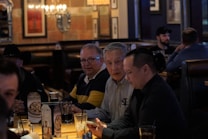 A group of people sitting around a wooden table in a dimly lit restaurant or bar setting. The focus is on three men engaged in conversation. There are drinks on the table, and the atmosphere appears casual and relaxed with a chandelier and framed art in the background.