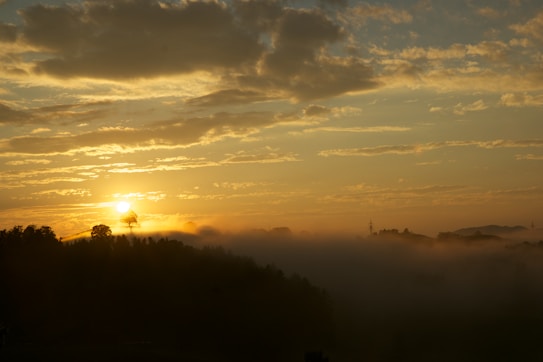 The image depicts a serene landscape with the sun setting or rising over a forested area. The sky is filled with dispersing clouds, some of them catching the sunlight, creating a warm golden glow. The forest below is shrouded in a layer of mist, adding a mystical and tranquil atmosphere.