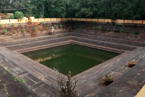 A stepped tank surrounded by stone steps, with green water at the bottom. The area around the tank features potted plants and greenery. The perimeter of the structure is outlined by a cream-colored wall with red accents. Trees can be seen in the background, adding to the natural setting.