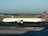 A Delta Airlines airplane is positioned on a tarmac, ready for takeoff or landing. The aircraft, a large commercial jet, is painted predominantly white with blue accents and a red and blue tail fin featuring Delta's logo. The runway and taxiway markings are visible, and the scene is set against a backdrop of an expansive airport area with clear skies and distant trees.