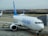 An Air Europa airplane is parked on the airport tarmac, connected to a jet bridge. The aircraft has a white body and blue tail, with the airline's logo prominently displayed. Other planes are visible in the background, along with a landscape of hills and trees.