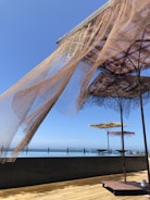 Large shade structures with net-like fabric are situated on a wooden deck overlooking a glassy, calm body of water. The sky is clear and blue, indicating a sunny day. The fabric of the shades is partially draped and flows naturally with the breeze, creating an airy and relaxed atmosphere.