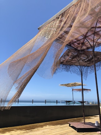 Large shade structures with net-like fabric are situated on a wooden deck overlooking a glassy, calm body of water. The sky is clear and blue, indicating a sunny day. The fabric of the shades is partially draped and flows naturally with the breeze, creating an airy and relaxed atmosphere.