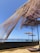 Large shade structures with net-like fabric are situated on a wooden deck overlooking a glassy, calm body of water. The sky is clear and blue, indicating a sunny day. The fabric of the shades is partially draped and flows naturally with the breeze, creating an airy and relaxed atmosphere.