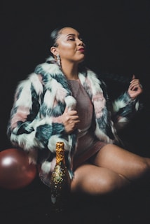 A person wearing a stylish, multicolored fur coat, paired with large, elegant earrings. They are sitting with a bottle decorated with golden sparkles in front of them, and a pink balloon partially visible. The background is dark, enhancing the dramatic and glamorous appearance.