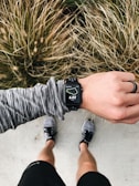 An overhead view of a person wearing running gear, looking at a smartwatch displaying 6.02 miles on a running route. The person is standing on a concrete surface with grassy foliage in the background. The individual is wearing a long-sleeved, gray-patterned top, black shorts, and running shoes. A black ring is worn on their left hand.