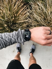 An overhead view of a person wearing running gear, looking at a smartwatch displaying 6.02 miles on a running route. The person is standing on a concrete surface with grassy foliage in the background. The individual is wearing a long-sleeved, gray-patterned top, black shorts, and running shoes. A black ring is worn on their left hand.