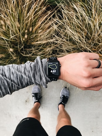 An overhead view of a person wearing running gear, looking at a smartwatch displaying 6.02 miles on a running route. The person is standing on a concrete surface with grassy foliage in the background. The individual is wearing a long-sleeved, gray-patterned top, black shorts, and running shoes. A black ring is worn on their left hand.