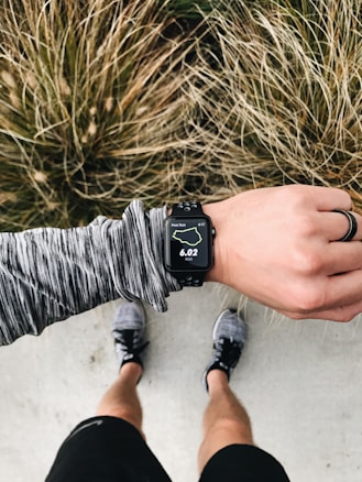An overhead view of a person wearing running gear, looking at a smartwatch displaying 6.02 miles on a running route. The person is standing on a concrete surface with grassy foliage in the background. The individual is wearing a long-sleeved, gray-patterned top, black shorts, and running shoes. A black ring is worn on their left hand.