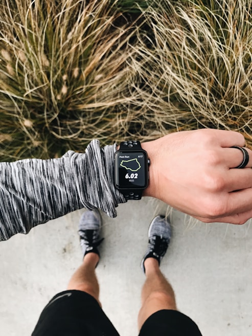 An overhead view of a person wearing running gear, looking at a smartwatch displaying 6.02 miles on a running route. The person is standing on a concrete surface with grassy foliage in the background. The individual is wearing a long-sleeved, gray-patterned top, black shorts, and running shoes. A black ring is worn on their left hand.