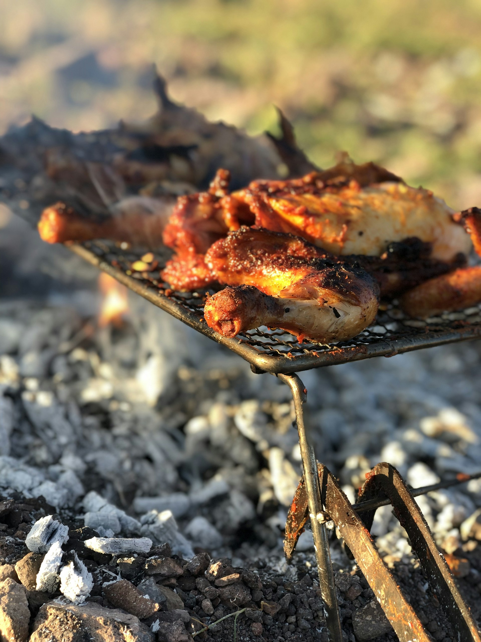 Grilled or roasted meat, likely chicken, with a crispy texture, placed on a grill over charcoal with visible ashes and embers. The setting suggests outdoor cooking, possibly a barbecue.
