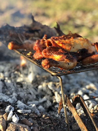 Grilled or roasted meat, likely chicken, with a crispy texture, placed on a grill over charcoal with visible ashes and embers. The setting suggests outdoor cooking, possibly a barbecue.