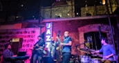 A jazz band is performing live in a cozy, dimly lit venue with brick walls and large windows. The group consists of a pianist, guitarist, bassist, saxophonist, and drummer. A neon sign reading 'JAZZ' is illuminated in blue light. The setting creates an intimate and vibrant atmosphere with soft lighting effects.