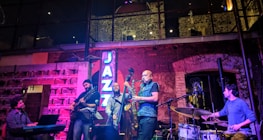 A jazz band is performing live in a cozy, dimly lit venue with brick walls and large windows. The group consists of a pianist, guitarist, bassist, saxophonist, and drummer. A neon sign reading 'JAZZ' is illuminated in blue light. The setting creates an intimate and vibrant atmosphere with soft lighting effects.