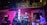 A jazz band is performing live in a cozy, dimly lit venue with brick walls and large windows. The group consists of a pianist, guitarist, bassist, saxophonist, and drummer. A neon sign reading 'JAZZ' is illuminated in blue light. The setting creates an intimate and vibrant atmosphere with soft lighting effects.