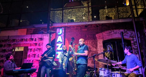 A jazz band is performing live in a cozy, dimly lit venue with brick walls and large windows. The group consists of a pianist, guitarist, bassist, saxophonist, and drummer. A neon sign reading 'JAZZ' is illuminated in blue light. The setting creates an intimate and vibrant atmosphere with soft lighting effects.