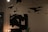 A dimly lit room features modern art hanging from the ceiling, including abstract paper airplanes. An abstract black and white painting hangs on the wall, alongside a lamp with exposed bulbs and a green decorative object on a table.