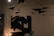 A dimly lit room features modern art hanging from the ceiling, including abstract paper airplanes. An abstract black and white painting hangs on the wall, alongside a lamp with exposed bulbs and a green decorative object on a table.