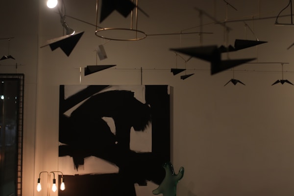 A dimly lit room features modern art hanging from the ceiling, including abstract paper airplanes. An abstract black and white painting hangs on the wall, alongside a lamp with exposed bulbs and a green decorative object on a table.