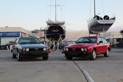 Two classic cars are parked in an industrial area near a boatyard, where two yachts are elevated on stilts. The setting appears to be at a boat maintenance facility with concrete ground.