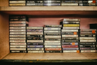 A vintage collection of music cassette tapes is organized on a wooden shelf. The tapes are stacked in groups and feature various artists and bands, each with distinct album covers and titles. The shelf has a rustic appearance with hints of wear and age, adding a nostalgic feel.