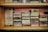 A vintage collection of music cassette tapes is organized on a wooden shelf. The tapes are stacked in groups and feature various artists and bands, each with distinct album covers and titles. The shelf has a rustic appearance with hints of wear and age, adding a nostalgic feel.