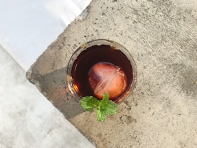 A glass of dark brown iced beverage with a large ice cube and a sprig of fresh mint is placed on a textured stone surface. The lighting creates a contrast between the cool drink and the rough, light-colored texture of the surface.